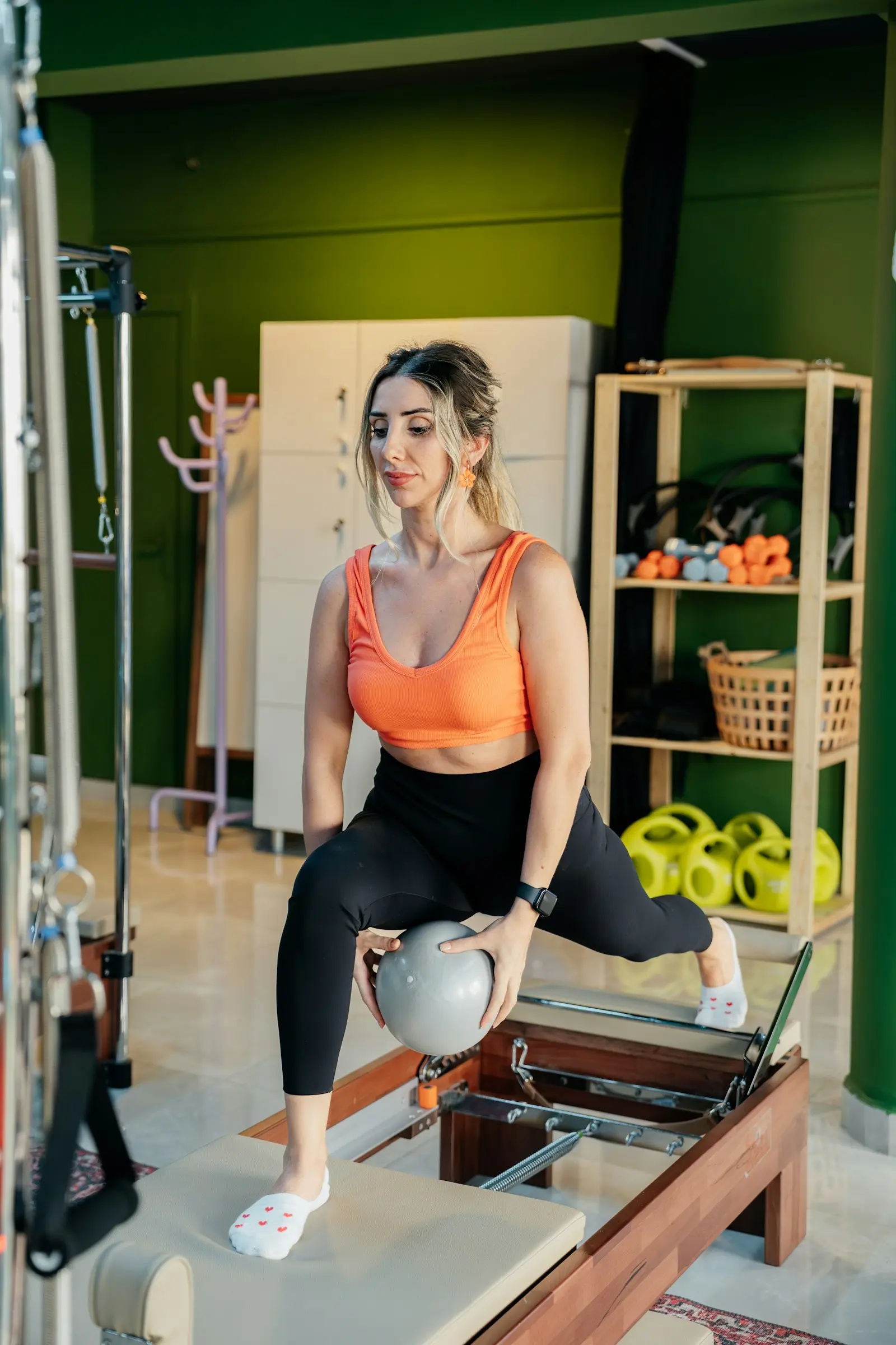 Femme en tenue orange qui fait un exercice avec un ballon sur un Reformer