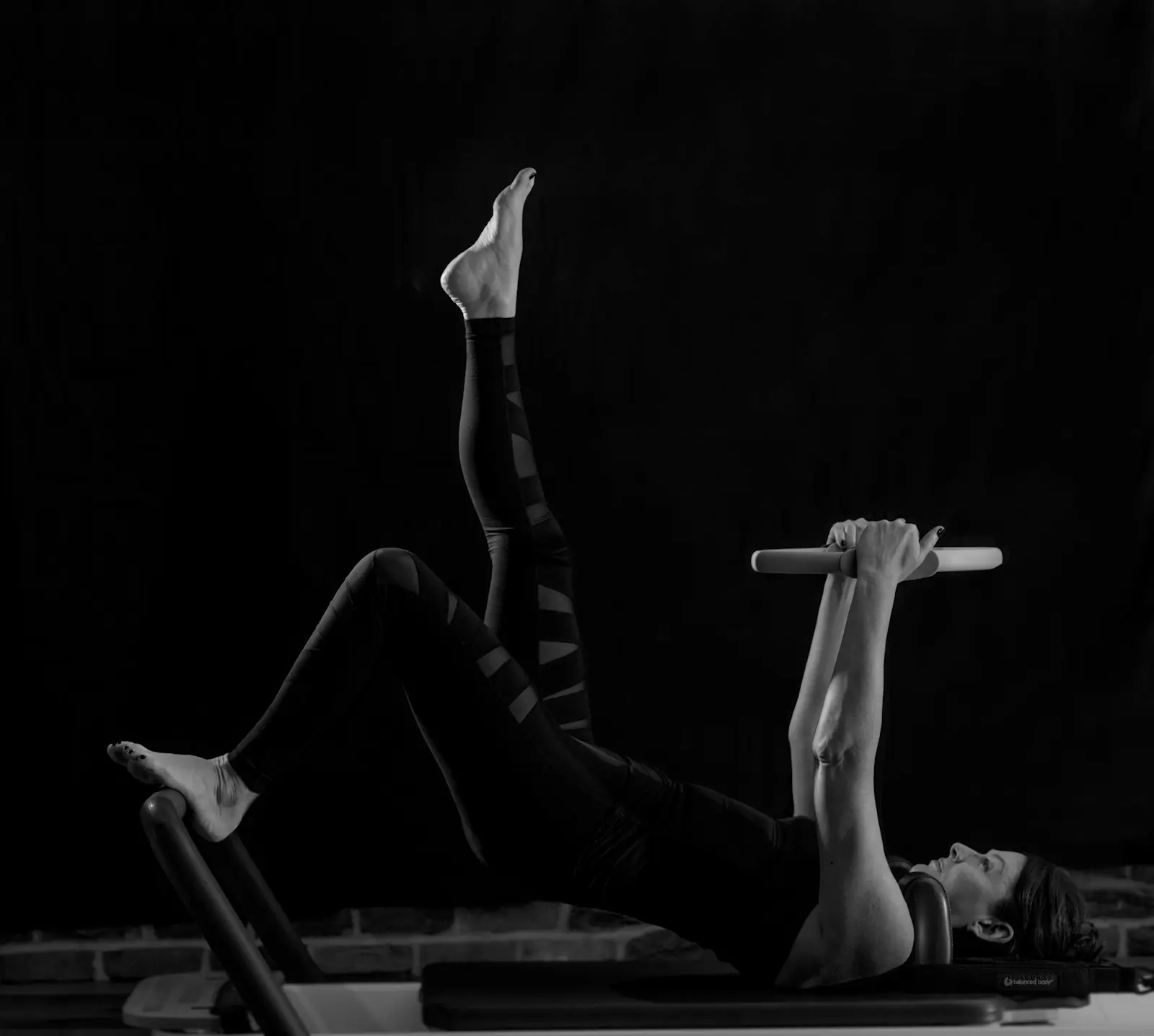 Femme allongée, jambes levées, qui tient une barre, sur fond noir